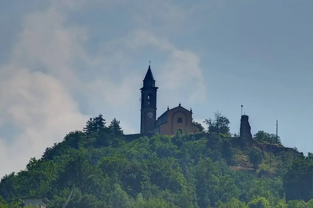 Chiesa e ruderi di un castello su una collina verdeggiante a Santa Margherita di Staffora, sotto un cielo nuvoloso.