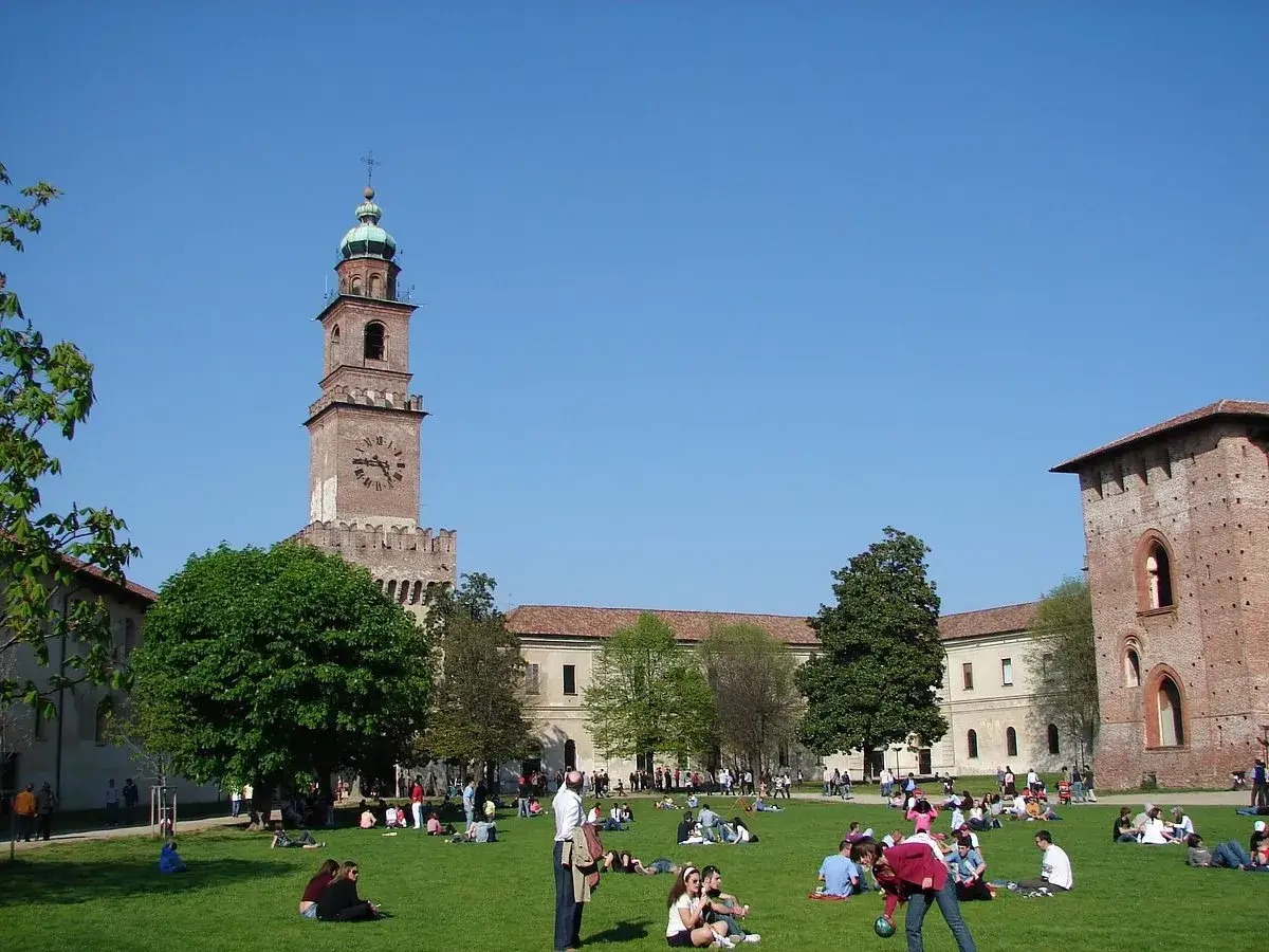 La piazza del Castello Sforzesco a Vigevano, con la torre dell'orologio e gente che si rilassa sull'erba. Un luogo ideale per scoprire cosa vedere a Vigevano.