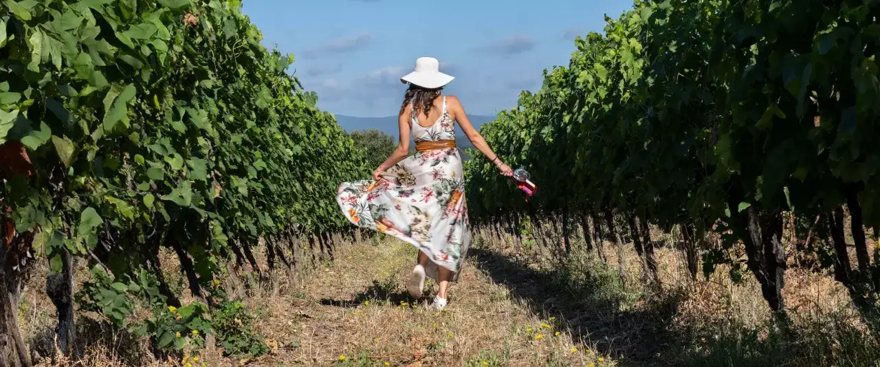 Donna con cappello bianco e abito fiorato cammina tra i filari di viti, tenendo in mano una bottiglia di vino e un calice, forse un Brunello di Montalcino.