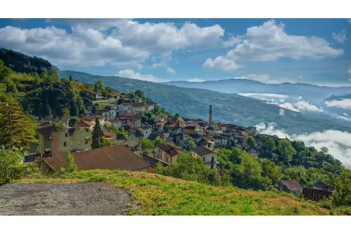 Un borgo di Pregola aggrappato alla collina, con case colorate e un campanile che svetta tra il verde. Nuvole basse accarezzano le montagne sullo sfondo.
