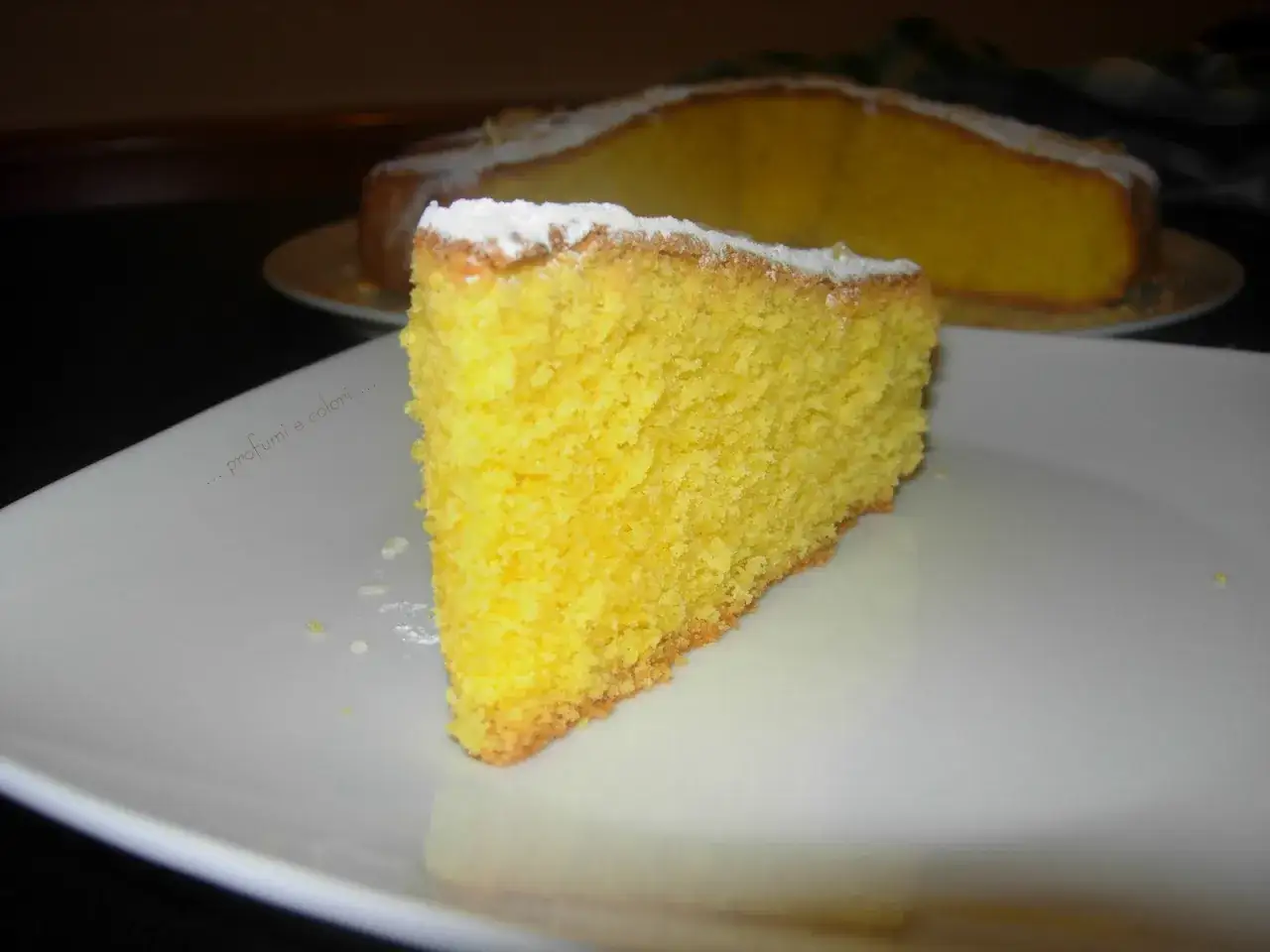 Una fetta soffice di torta di mandorle, cosparsa di zucchero a velo, su un piatto bianco.