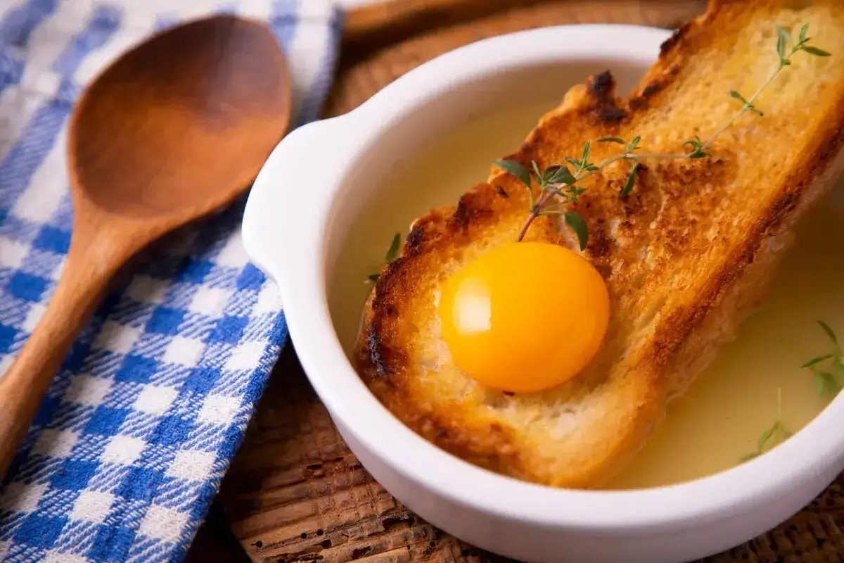 Zuppa alla pavese in una ciotola bianca, con pane tostato, tuorlo d'uovo e timo. Accanto, un cucchiaio di legno e un tovagliolo a quadri blu.