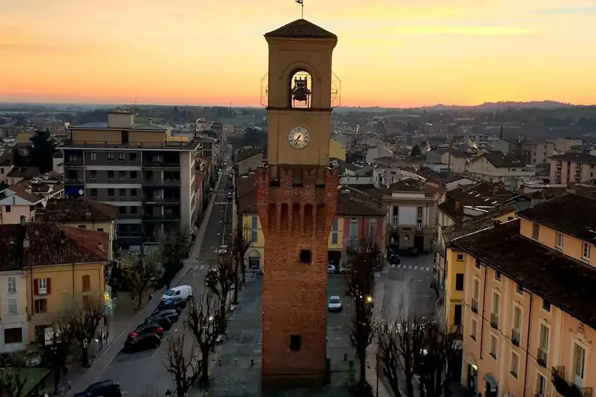 Torre dell'orologio in una piazza alberata, con case colorate e un tramonto dorato. Una stradella da esplorare, con tanto da vedere.