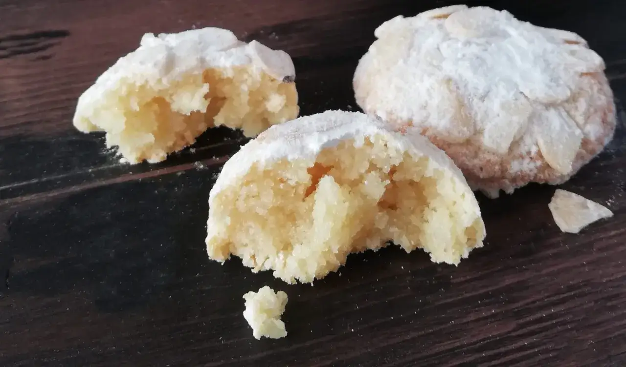 Biscotti ovali cosparsi di zucchero a velo e mandorle a lamelle, uno &egrave; spezzato mostrando l'interno friabile.