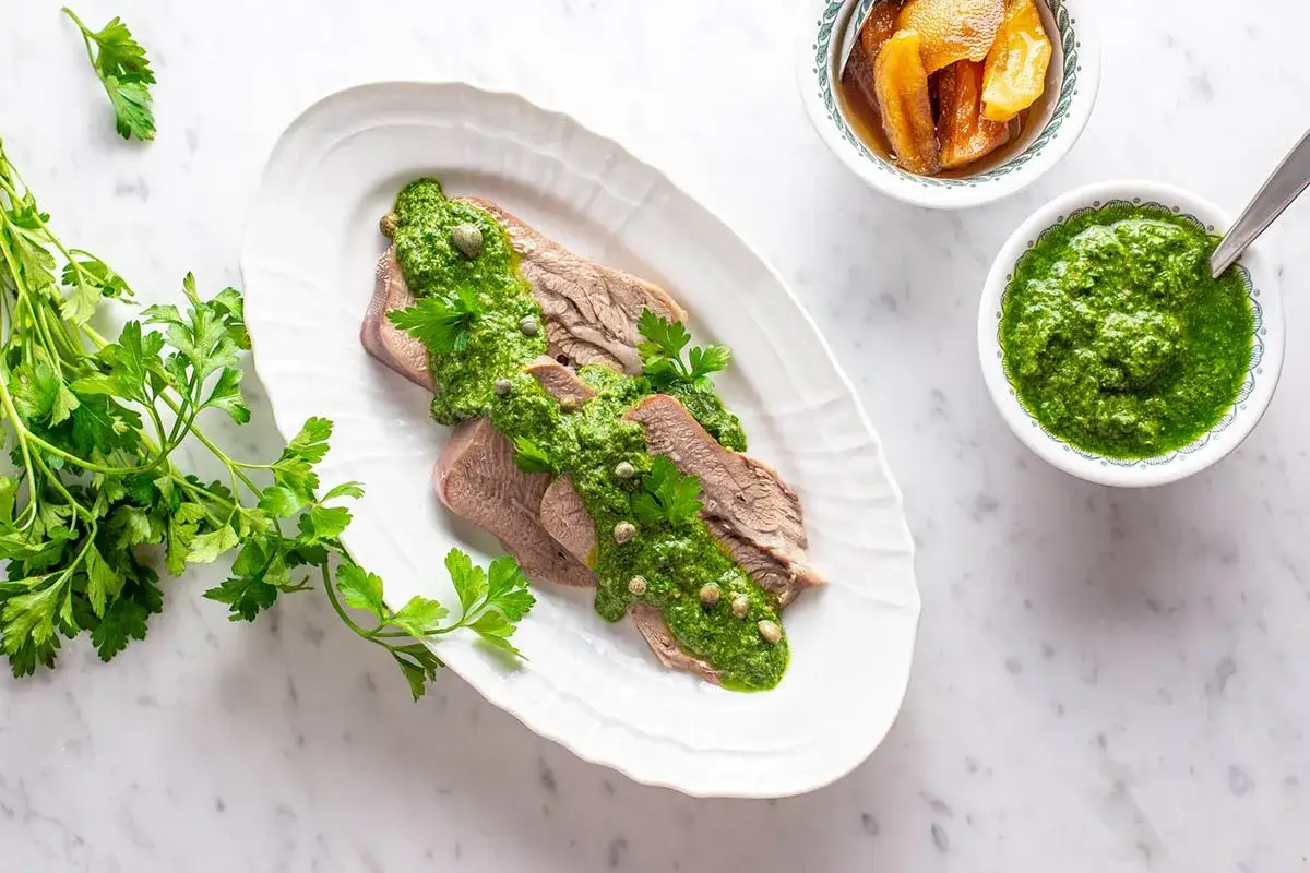 Lingua bollita affettata, generosamente condita con salsa verde e capperi. Accanto, una ciotola di salsa verde e altra guarnizione.