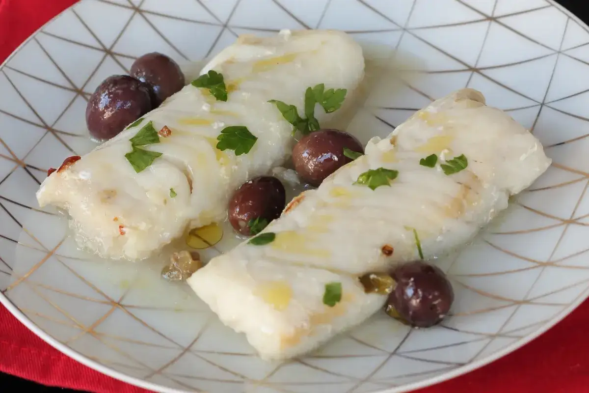 Merluzzo con cipolle in bianco, guarnito con olive nere e prezzemolo fresco, su un piatto bianco con decorazioni dorate.