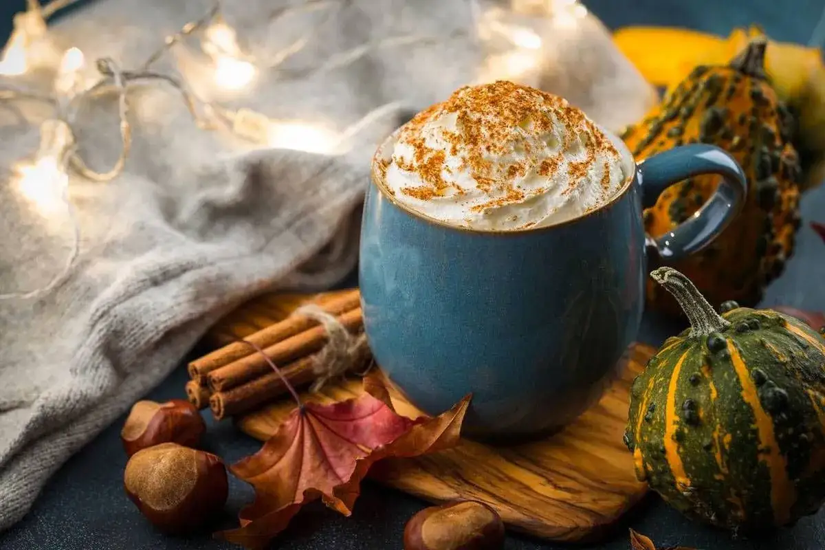 Tazza blu con panna montata e cannella, circondata da castagne, foglie autunnali, bastoncini di cannella e piccole zucche. Un'atmosfera calda per gustare castagne al latte.