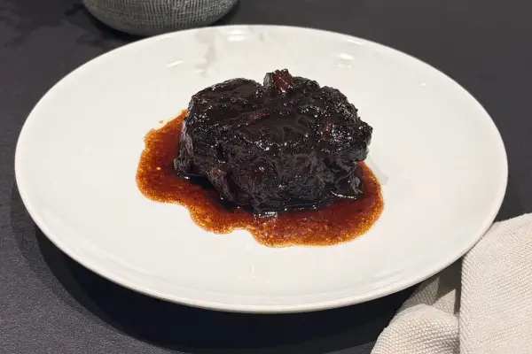 Un succulento brasato al buttafuoco, lucido e scuro, adagiato su un piatto bianco con un sugo denso e aromatico.