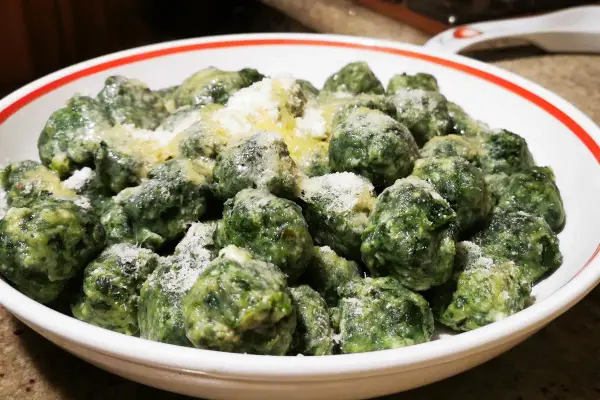 Malfatti ricetta originale: gnocchetti verdi fatti in casa, conditi con burro fuso e parmigiano grattugiato, serviti in una ciotola bianca con bordo rosso.